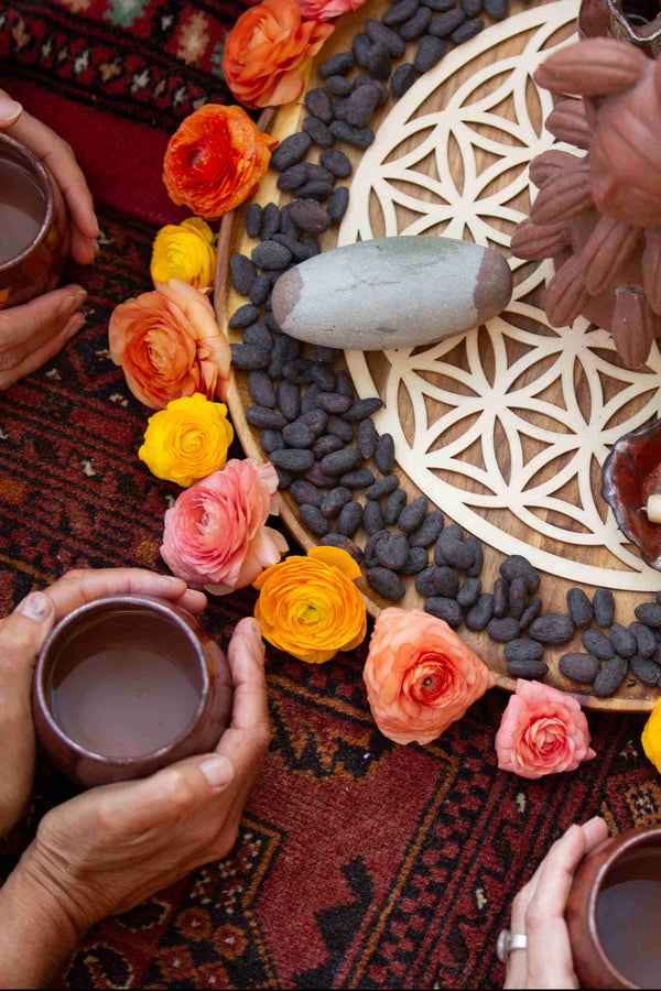 People taking part in a sacred cacao ceremony , paying reverence to the cacao goddess who sits atop of a central altar surrounded by chuncho cacao beans, stunning roses, a shiva lingum, al supported by a flower of life sacred geometric symbol.