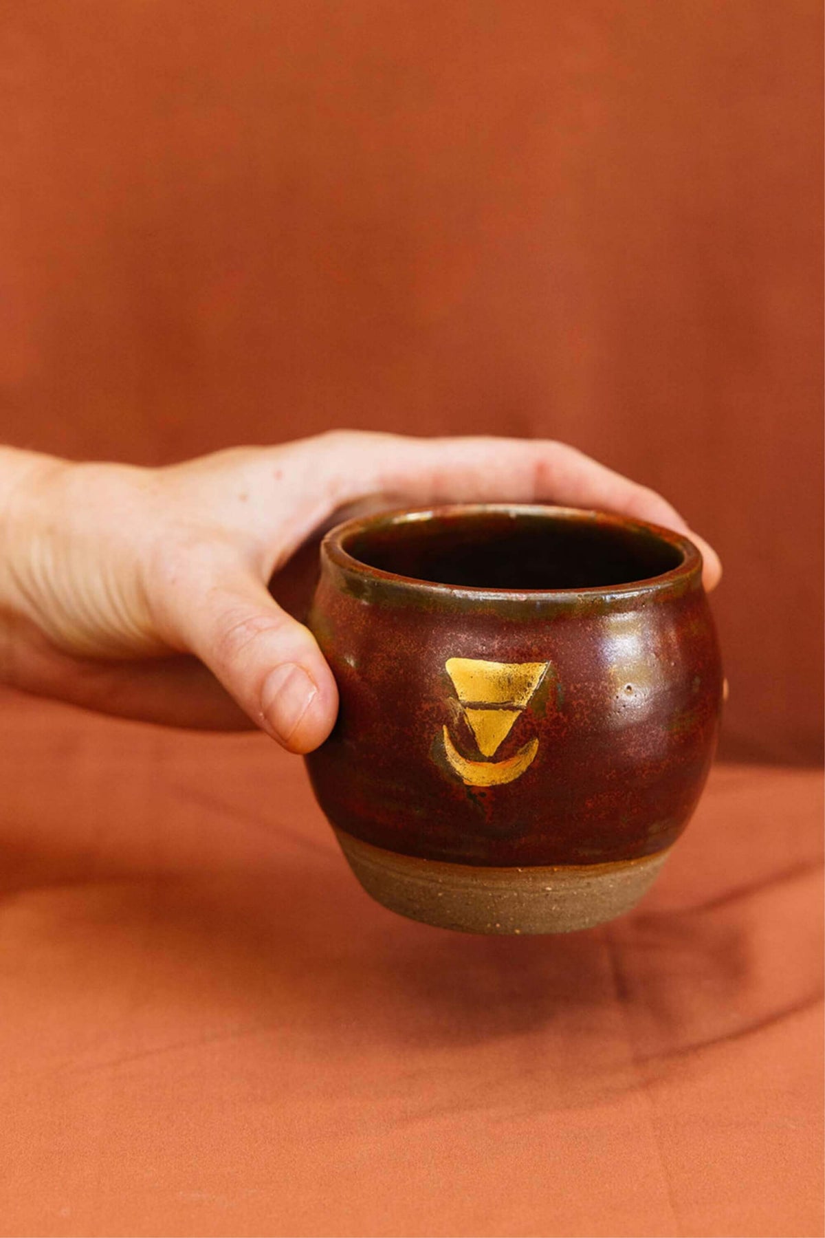 Hand holding a small ceremonial cup, embossed with a real gold lustre Sacred Earth Medicine symbol. The cup is made from wild harvested clay sourced using methods taught by the indigenous people from the Northern Rivers of NSW.